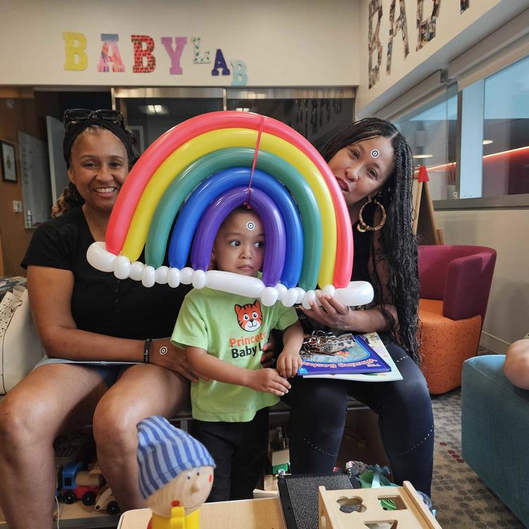Mother and grandmother at the Baby Lab hold a rainbow made of balloons around a child's face