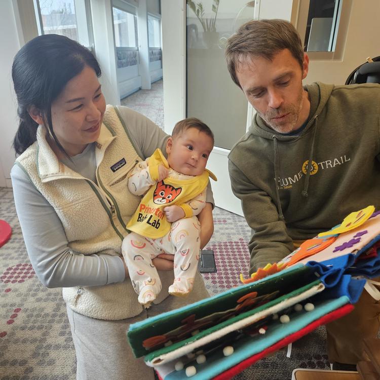 A mother holds her baby who is wearing a baby lab bib while a researcher reads a book to the baby